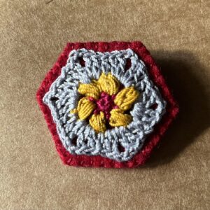 A hexagon shaped, hand stitched, felt and flower design crocheted brooch with metal locking brooch fixing.
