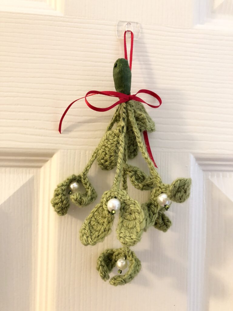 A handmade, crocheted, hanging mistletoe decoration. Made using green yarn and glass beads which are grouped and wired together and tied with a red ribbon to hang. small size approximately 22cm high (not including ribbon hanging loop.) 5 springs of mistletoe are used in this arrangement.