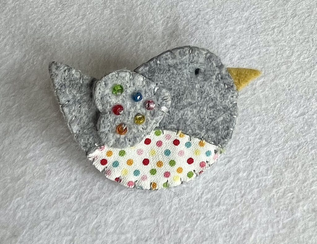 This little birdie brooch is completely hand cut, stitched embroidered and embellished. It is made using a wool mix felt and patterned fabric. It is hand embellished using glass beads and has a metal locking fixing on the back. Approximate size 6cm wide x 4cm height. 