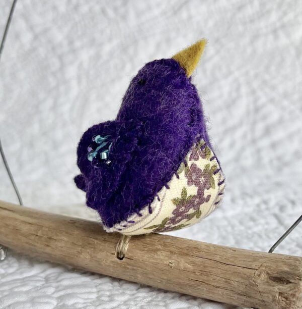 A single small sized bird, handmade in purple coloured felt, with a cotton purple flower print fabric chest and hand embroidered/glass beaded detail on the wings. This bird is sat on a natural driftwood perch with a wire hanger that is decorated with a crocheted flower and leaf detail.