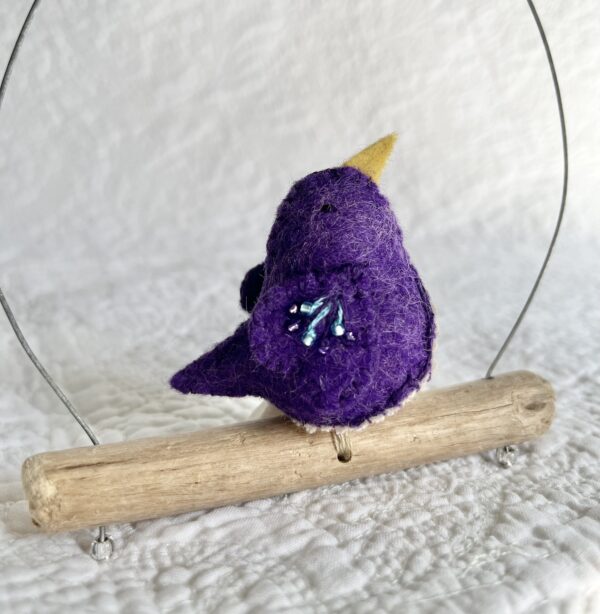 A single small sized bird, handmade in purple coloured felt, with a cotton purple flower print fabric chest and hand embroidered/glass beaded detail on the wings. This bird is sat on a natural driftwood perch with a wire hanger that is decorated with a crocheted flower and leaf detail.