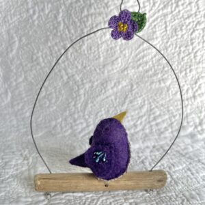 A single small sized bird, handmade in purple coloured felt, with a cotton purple flower print fabric chest and hand embroidered/glass beaded detail on the wings. This bird is sat on a natural driftwood perch with a wire hanger that is decorated with a crocheted flower and leaf detail.