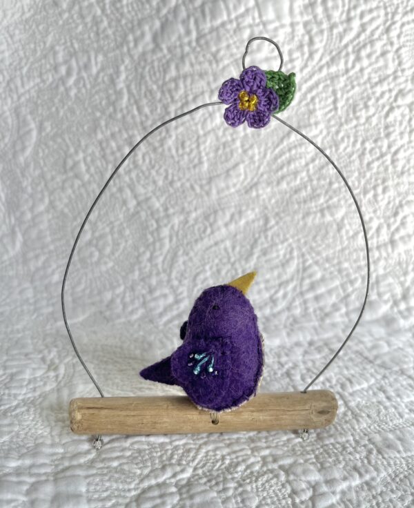 A single small sized bird, handmade in purple coloured felt, with a cotton purple flower print fabric chest and hand embroidered/glass beaded detail on the wings. This bird is sat on a natural driftwood perch with a wire hanger that is decorated with a crocheted flower and leaf detail.