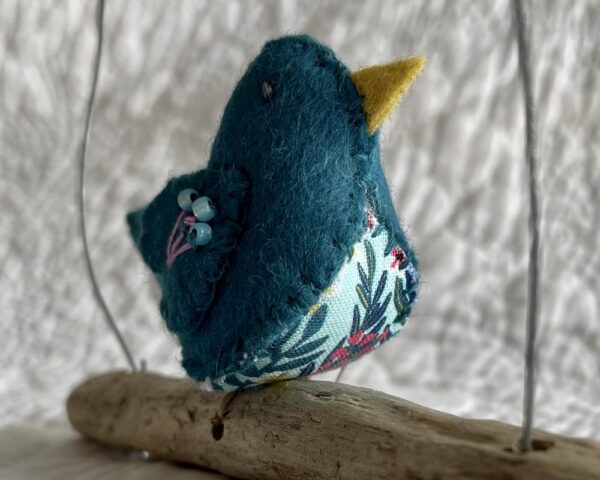 A single small sized bird, handmade in Teal coloured felt, with a cotton multi coloured flower print fabric chest and hand embroidered/glass beaded detail on the wings. This bird is sat on a natural driftwood perch with a wire hanger that is decorated with a crocheted pink flower detail.