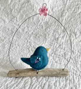 A single small sized bird, handmade in Teal coloured felt, with a cotton multi coloured flower print fabric chest and hand embroidered/glass beaded detail on the wings. This bird is sat on a natural driftwood perch with a wire hanger that is decorated with a crocheted pink flower detail.