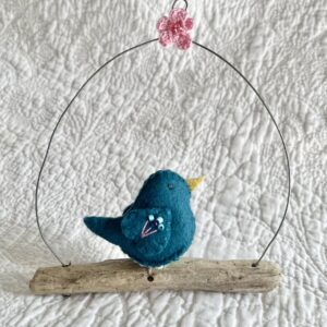 A single small sized bird, handmade in Teal coloured felt, with a cotton multi coloured flower print fabric chest and hand embroidered/glass beaded detail on the wings. This bird is sat on a natural driftwood perch with a wire hanger that is decorated with a crocheted pink flower detail.