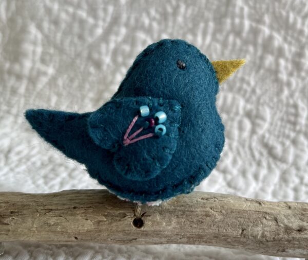 A single small sized bird, handmade in Teal coloured felt, with a cotton multi coloured flower print fabric chest and hand embroidered/glass beaded detail on the wings. This bird is sat on a natural driftwood perch with a wire hanger that is decorated with a crocheted pink flower detail.