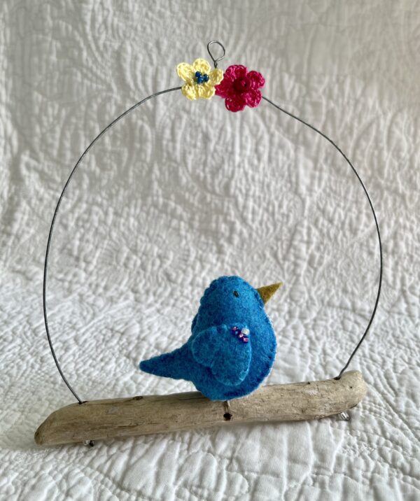 A single small sized bird, handmade in Turquoise coloured felt, with a cotton multi coloured dotty print fabric chest and hand embroidered/glass beaded detail on the wings. This bird is sat on a natural driftwood perch with a wire hanger that is decorated with a yellow and pink crocheted flowers.