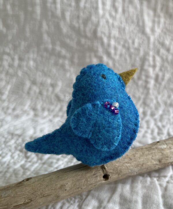 A single small sized bird, handmade in Turquoise coloured felt, with a cotton multi coloured dotty print fabric chest and hand embroidered/glass beaded detail on the wings. This bird is sat on a natural driftwood perch with a wire hanger that is decorated with a yellow and pink crocheted flowers.