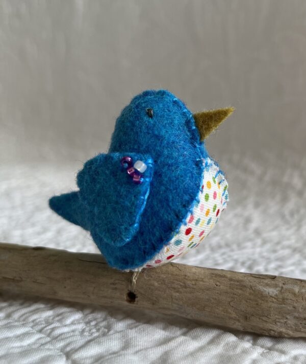 A single small sized bird, handmade in Turquoise coloured felt, with a cotton multi coloured dotty print fabric chest and hand embroidered/glass beaded detail on the wings. This bird is sat on a natural driftwood perch with a wire hanger that is decorated with a yellow and pink crocheted flowers.