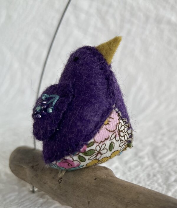 A single small sized bird, handmade in purple coloured felt, with a cotton purple flower print fabric chest and hand embroidered/glass beaded detail on the wings. This bird is sat on a natural driftwood perch with a wire hanger that is decorated with a crocheted flower and leaf detail.