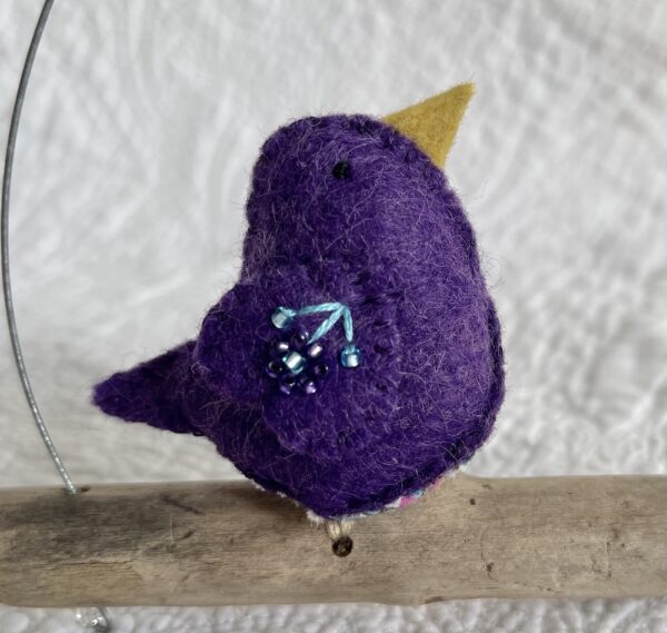 A single small sized bird, handmade in purple coloured felt, with a cotton purple flower print fabric chest and hand embroidered/glass beaded detail on the wings. This bird is sat on a natural driftwood perch with a wire hanger that is decorated with a crocheted flower and leaf detail.