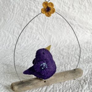 A single small sized bird, handmade in purple coloured felt, with a cotton purple flower print fabric chest and hand embroidered/glass beaded detail on the wings. This bird is sat on a natural driftwood perch with a wire hanger that is decorated with a crocheted flower and leaf detail.