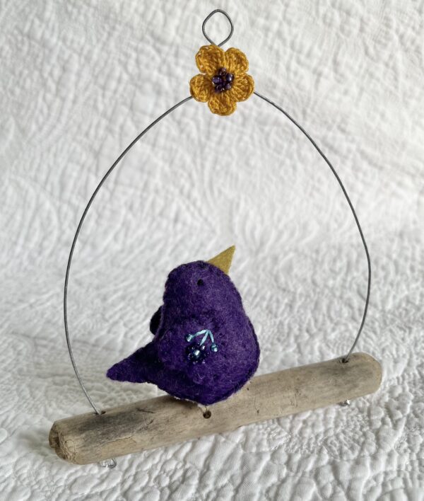 A single small sized bird, handmade in purple coloured felt, with a cotton purple flower print fabric chest and hand embroidered/glass beaded detail on the wings. This bird is sat on a natural driftwood perch with a wire hanger that is decorated with a crocheted flower and leaf detail.