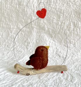 A single, small sized bird, handmade in brown felt with a red and white spotty cotton fabric chest. The bird is sat on a natural driftwood perch with a wire hanger that is decorated with a single red heart. Approximate size 11cm width x 14cm 