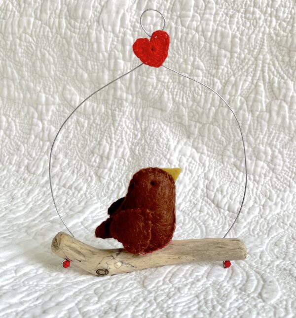 A single, small sized bird, handmade in brown felt with a red and white spotty cotton fabric chest. The bird is sat on a natural driftwood perch with a wire hanger that is decorated with a single red heart. Approximate size 11cm width x 14cm