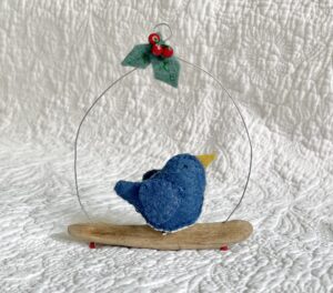 A single, small sized bird, handmade in denim blue felt with a cotton holly print fabric chest. The bird is sat on a natural driftwood perch with a wire hanger that is decorated with felt holly leaves and glass beads. Approximate size 12cm width x 14cm height.