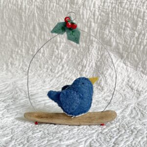A single, small sized bird, handmade in denim blue felt with a cotton holly print fabric chest. The bird is sat on a natural driftwood perch with a wire hanger that is decorated with felt holly leaves and glass beads. Approximate size 12cm width x 14cm height.