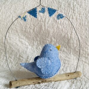 A medium sized bird, handmade in light blue coloured felt, with a cotton forget-me-not print fabric chest. This bird is sat on a natural driftwood perch with a wire hanger that is decorated with coordinating mini bunting.