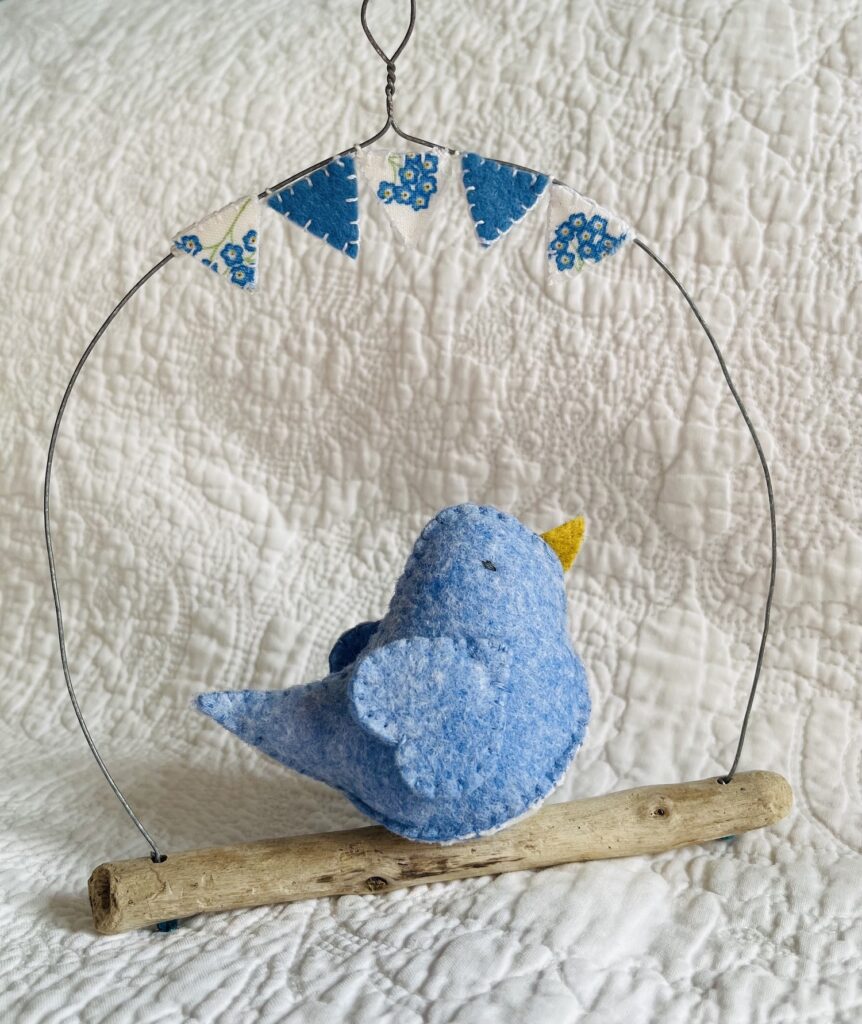 A medium sized bird, handmade in light blue coloured felt, with a cotton forget-me-not print fabric chest. This bird is sat on a natural driftwood perch with a wire hanger that is decorated with coordinating mini bunting.