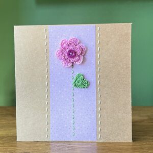 A single crocheted flower with a double layer of Lilac petals and button, with a green leaf and hand stitched detailing. This medium brown card has a blank white insert for your own message, and an envelope is included. Approximate size 12.5cm x 12.5 cm. Made using 100% cotton. Eco-friendly, Vegan friendly and fully recyclable.