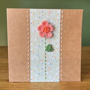A single crocheted peach/coral flower and button, with a green leaf and hand stitched detailing. This medium brown card has a blank white insert for your own message, and an envelope is included. Approximate size 12.5cm x 12.5 cm. Made using 100% cotton. Eco-friendly, Vegan friendly and fully recyclable.