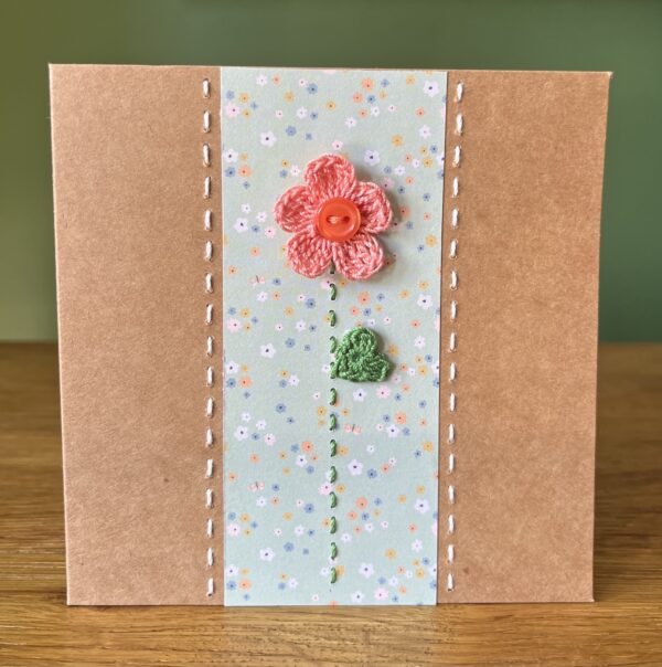A single crocheted peach/coral flower and button, with a green leaf and hand stitched detailing. This medium brown card has a blank white insert for your own message, and an envelope is included. Approximate size 12.5cm x 12.5 cm. Made using 100% cotton. Eco-friendly, Vegan friendly and fully recyclable.