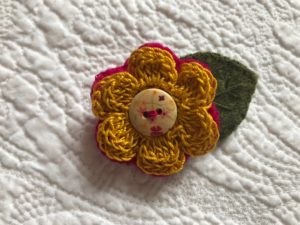 A crocheted mustard coloured cotton flower and green embroidered felt leaf brooch with decorative button detail.