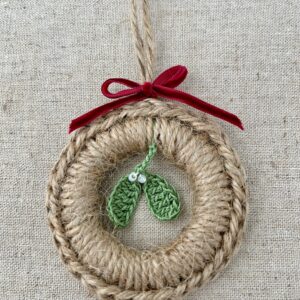 A crocheted and mini mistletoe embellished Christmas wreath, hanging decoration. Handmade using a wooden ring with 100% natural coloured Jute, 100% cotton in green and opal effect glass beaded embellishments. Approximate size of wreath 8.5cm height x 7cm width. Not including hanging loop.