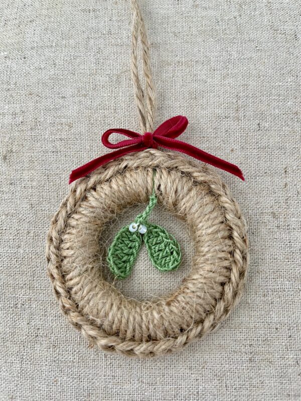A crocheted and mini mistletoe embellished Christmas wreath, hanging decoration. Handmade using a wooden ring with 100% natural coloured Jute, 100% cotton in green and opal effect glass beaded embellishments. Approximate size of wreath 8.5cm height x 7cm width. Not including hanging loop.