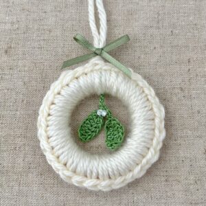 A crocheted and mini mistletoe embellished Christmas wreath, hanging decoration. Handmade using a wooden ring with cream coloured yarn, 100% cotton in green and opal effect glass beaded embellishments. Approximate size of wreath 8.5cm height x 7cm width. Not including hanging loop.