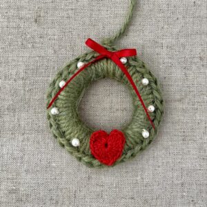 A crocheted and mini heart embellished Christmas wreath, hanging decoration. Handmade using a wooden ring with 100% green coloured Jute, 100% cotton red heart and pearl effect glass beaded embellishments, with a red bow. Approximate size of wreath 8.5cm height x 7cm width. Not including hanging loop.