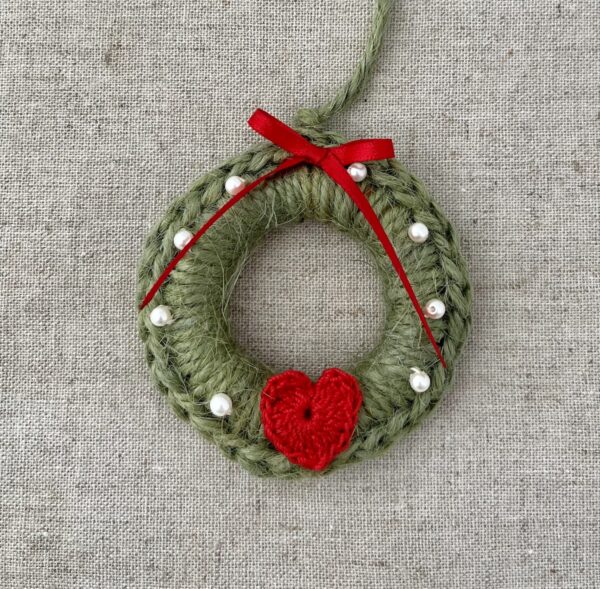 A crocheted and mini heart embellished Christmas wreath, hanging decoration. Handmade using a wooden ring with 100% green coloured Jute, 100% cotton red heart and pearl effect glass beaded embellishments, with a red bow. Approximate size of wreath 8.5cm height x 7cm width. Not including hanging loop.