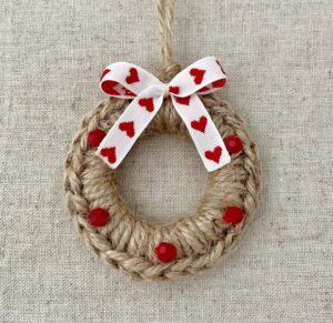 A crocheted and mini   embellished Christmas wreath, hanging decoration.

Handmade using a wooden ring with 100% natural coloured Jute and red glass beaded embellishments, with a heart detail bow.

Approximate size of wreath 8.5cm height x 7cm width. Not including hanging loop.