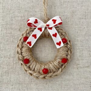 A crocheted and mini embellished Christmas wreath, hanging decoration. Handmade using a wooden ring with 100% natural coloured Jute and red glass beaded embellishments, with a heart detail bow. Approximate size of wreath 8.5cm height x 7cm width. Not including hanging loop.