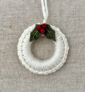 A crocheted and mini holly embellished Christmas wreath, hanging decoration.

Handmade using a wooden ring with cream coloured yarn, embellished with  green embroidered felt holly leaves and red glass beads.

Approximate size of wreath 8.5cm height x 7cm width. Not including hanging loop.
