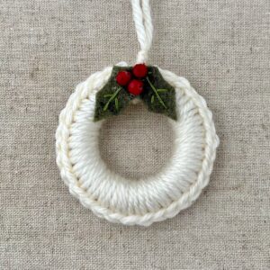 A crocheted and mini holly embellished Christmas wreath, hanging decoration. Handmade using a wooden ring with cream coloured yarn, embellished with green embroidered felt holly leaves and red glass beads. Approximate size of wreath 8.5cm height x 7cm width. Not including hanging loop.