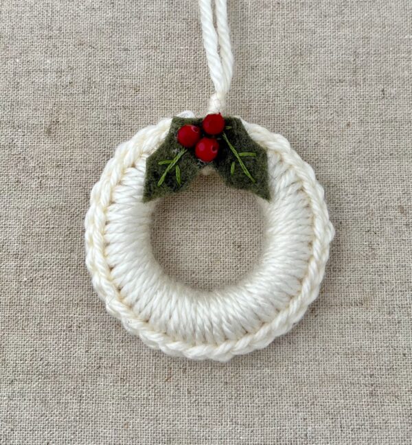 A crocheted and mini holly embellished Christmas wreath, hanging decoration. Handmade using a wooden ring with cream coloured yarn, embellished with green embroidered felt holly leaves and red glass beads. Approximate size of wreath 8.5cm height x 7cm width. Not including hanging loop.