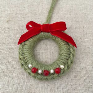 A crocheted and embellished Christmas wreath, hanging decoration. Handmade using a wooden ring with 100% green coloured Jute, red and pearl effect glass beaded embellishments, with a red velvet bow. Approximate size of wreath 8.5cm height x 7cm width. Not including hanging loop.