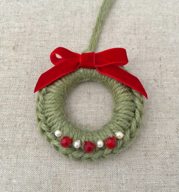 A crocheted and embellished Christmas wreath, hanging decoration. Handmade using a wooden ring with 100% green coloured Jute, red and pearl effect glass beaded embellishments, with a red velvet bow. Approximate size of wreath 8.5cm height x 7cm width. Not including hanging loop.