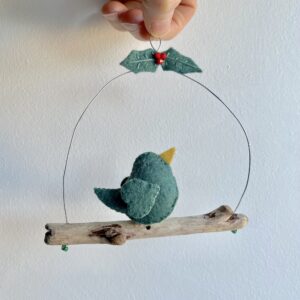 A single, small sized bird, handmade in green felt with a cotton holly print fabric chest. The bird is sat on a natural driftwood perch with a wire hanger that is decorated with felt holly leaves and glass beads. Approximate size 14cm width by 14cm height.