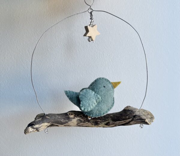 A single, small sized bird, handmade in duck egg green felt with a cotton mistletoe print fabric chest. The bird is sat on a natural driftwood perch with a wire hanger that is decorated with a wooden star and glass beads. Approximate size 14cm width x 15cm height.