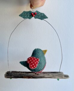A single, small sized bird, handmade in green felt with a red and white dotty cotton print fabric chest and wings.
The bird is sat on a natural driftwood perch with a wire hanger that is decorated with felt holly leaves and glass beads. Approximate size 13cm width by 15cm height.
