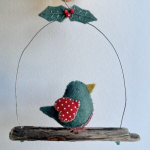 A single, small sized bird, handmade in green felt with a red and white dotty cotton print fabric chest and wings. The bird is sat on a natural driftwood perch with a wire hanger that is decorated with felt holly leaves and glass beads. Approximate size 13cm width by 15cm height.
