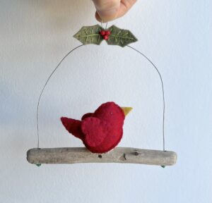 A single, small sized bird, handmade in red felt with a cotton holly print fabric chest. The bird is sat on a natural driftwood perch with a wire hanger that is decorated with  felt holly leaves and glass beads.

Approximate size 13cm width by 13cm height.