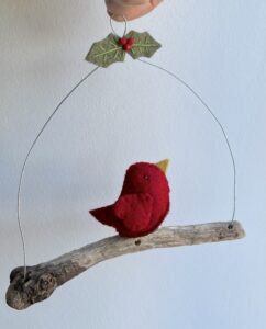 A single, small sized bird, handmade in red felt with a cotton holly print fabric chest. The bird is sat on a natural driftwood perch with a wire hanger that is decorated with felt holly leaves and glass beads. Approximate size 14cm width by 15cm height.