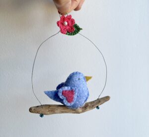 A single, small sized bird, handmade in Light blue felt with a cotton floral print fabric chest and pink heart detail on wings. The bird is sat on a natural driftwood perch with a wire hanger that is decorated with a crocheted flower and leaf and glass beads.

Approximate size 11cm width by 13cm height.
