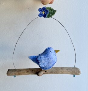A single, small sized bird, handmade in Light blue felt with a cotton forget-me-not print fabric chest. The bird is sat on a natural driftwood perch with a wire hanger that is decorated with a crocheted flower and leaf. Approximate size 15cm width by 15cm height.