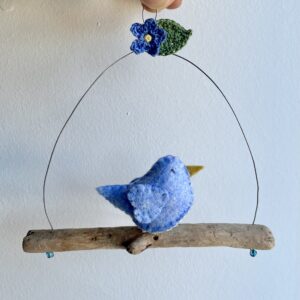 A single, small sized bird, handmade in Light blue felt with a cotton forget-me-not print fabric chest. The bird is sat on a natural driftwood perch with a wire hanger that is decorated with a crocheted flower and leaf. Approximate size 15cm width by 15cm height.