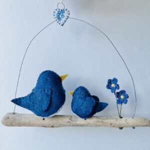 A Double bird decoration with one large and one small small sized bird, handmade in denim blue felt with a cotton forget-me-not print fabric chest. The birds are sat on a natural driftwood perch with a wire hanger that is decorated with a fabric heart and two crocheted flowers. Approximate size 22cm width by 20cm height.